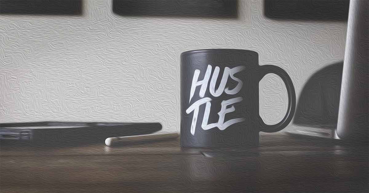 An image of a desk with a coffee mug on it. The mug has the word hustle printed on it.